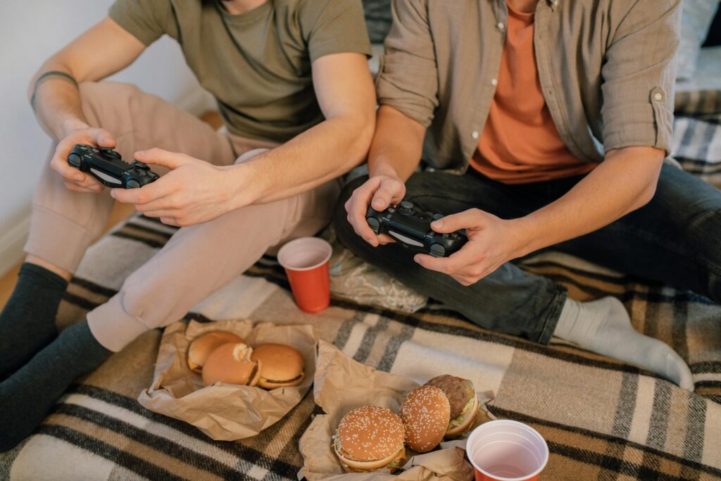 Two friends having fun playing video games with controllers and eating burgers indoors.