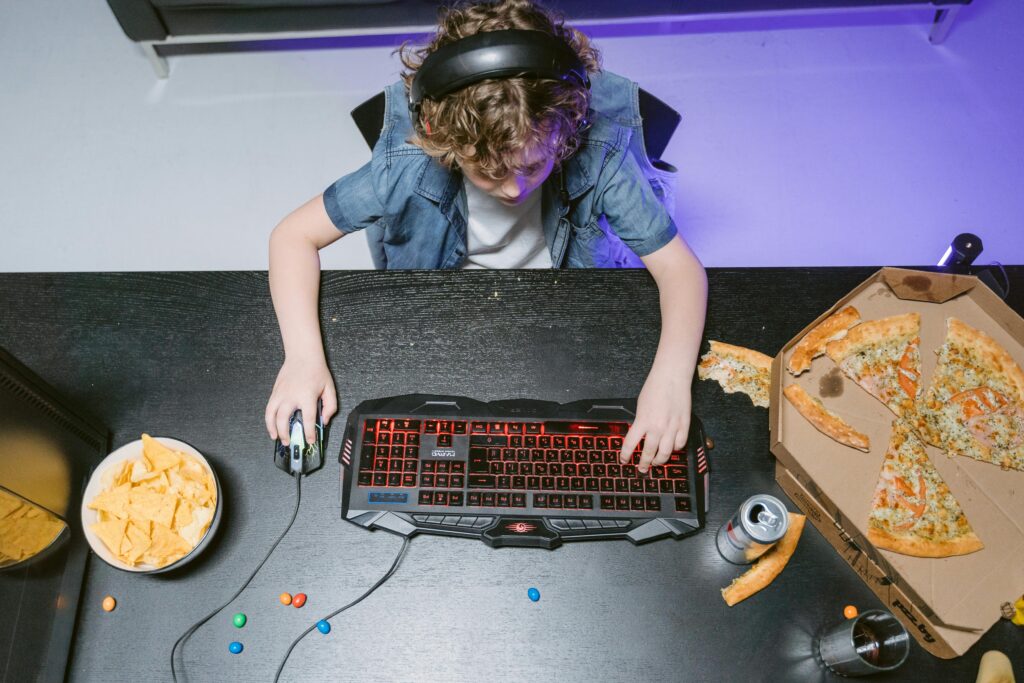 Top view of a child playing video games, surrounded by snacks like pizza and chips on a table.